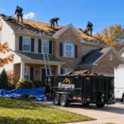 Empire Home Solutions crew tearing off old roofing on a two-story brick home, with a debris dumpster and work truck in the driveway.