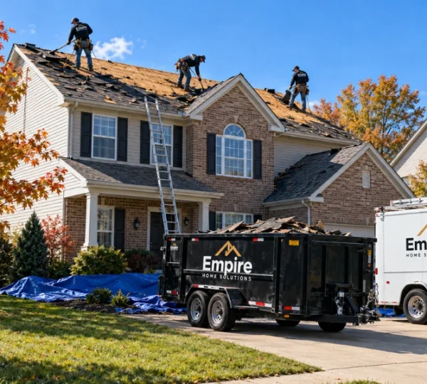 Empire Home Solutions crew tearing off old roofing on a two-story brick home, with a debris dumpster and work truck in the driveway.