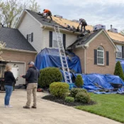 Roofers in safety harnesses installing underlayment and metal flashing beside a brick chimney during a roof replacement.
